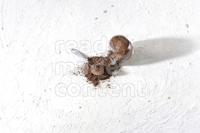 A flipped glass spice jar and a metal spoon full of cloves powder and powder came out of the jar on textured white flooring