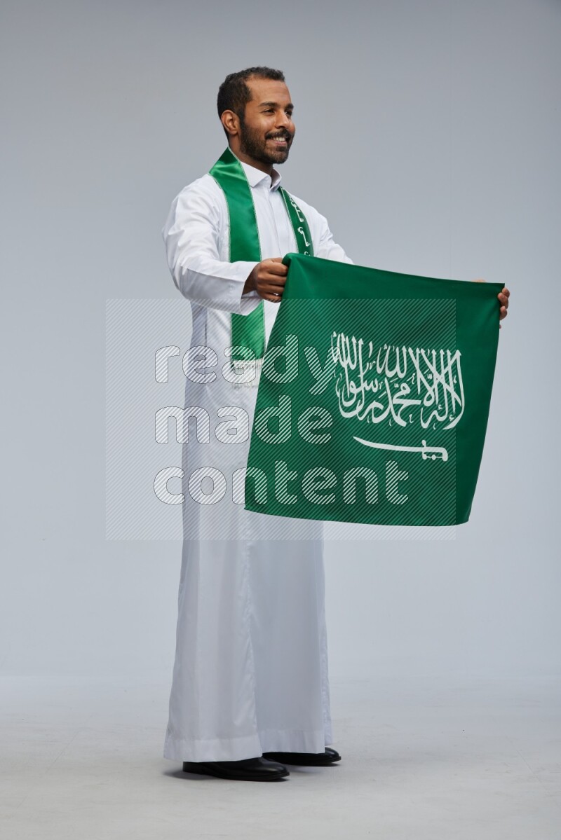 Saudi man wearing Thob standing holding Saudi flag on Gray background