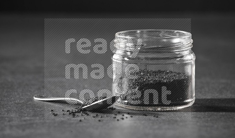A glass jar full of black seeds with a metal spoon full of the seeds on a black flooring