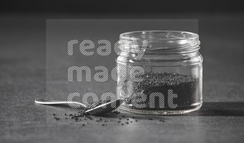 A glass jar full of black seeds with a metal spoon full of the seeds on a black flooring