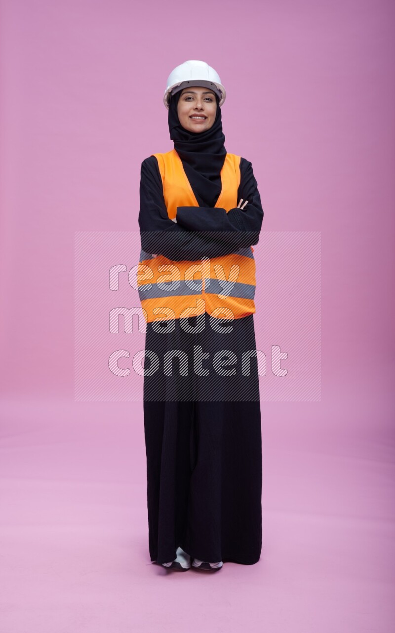 Saudi woman wearing Abaya with engineer vest and helmet standing with crossed arms on pink background