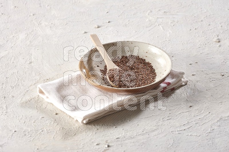 A multicolored pottery plate full of flax with a wooden spoon full of the seeds on a napkin on a textured white flooring in different angles