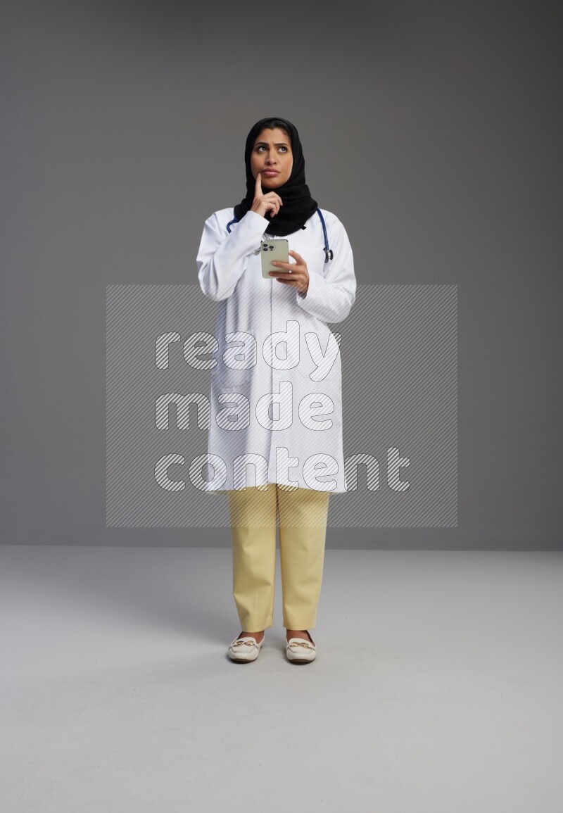 Saudi woman wearing lab coat with stethoscope standing texting on phone on Gray background
