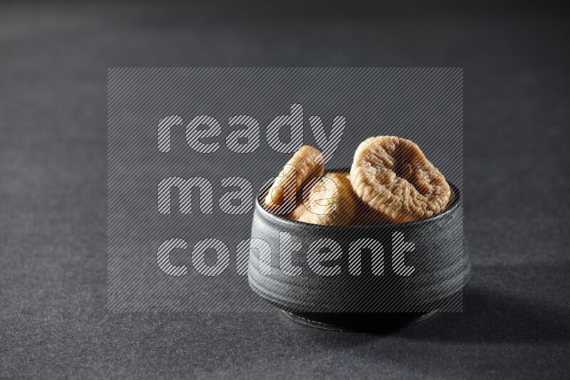 A black pottery bowl full of dried figs on a black background in different angles