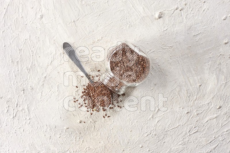 A glass spice jar full of flax seeds flipped with a metal spoon full of the seeds on a textured white flooring