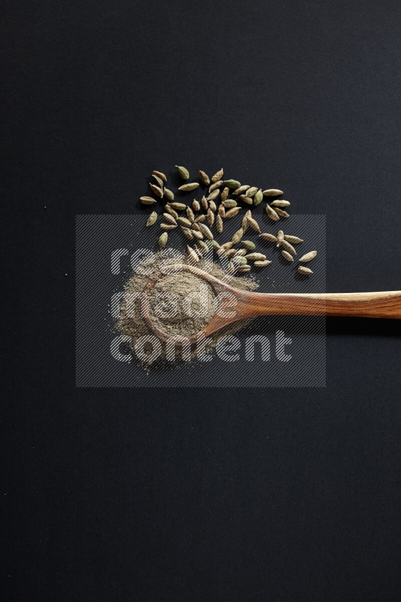 A wooden ladle full of cardamom powder and cardamom seeds beside it on black flooring