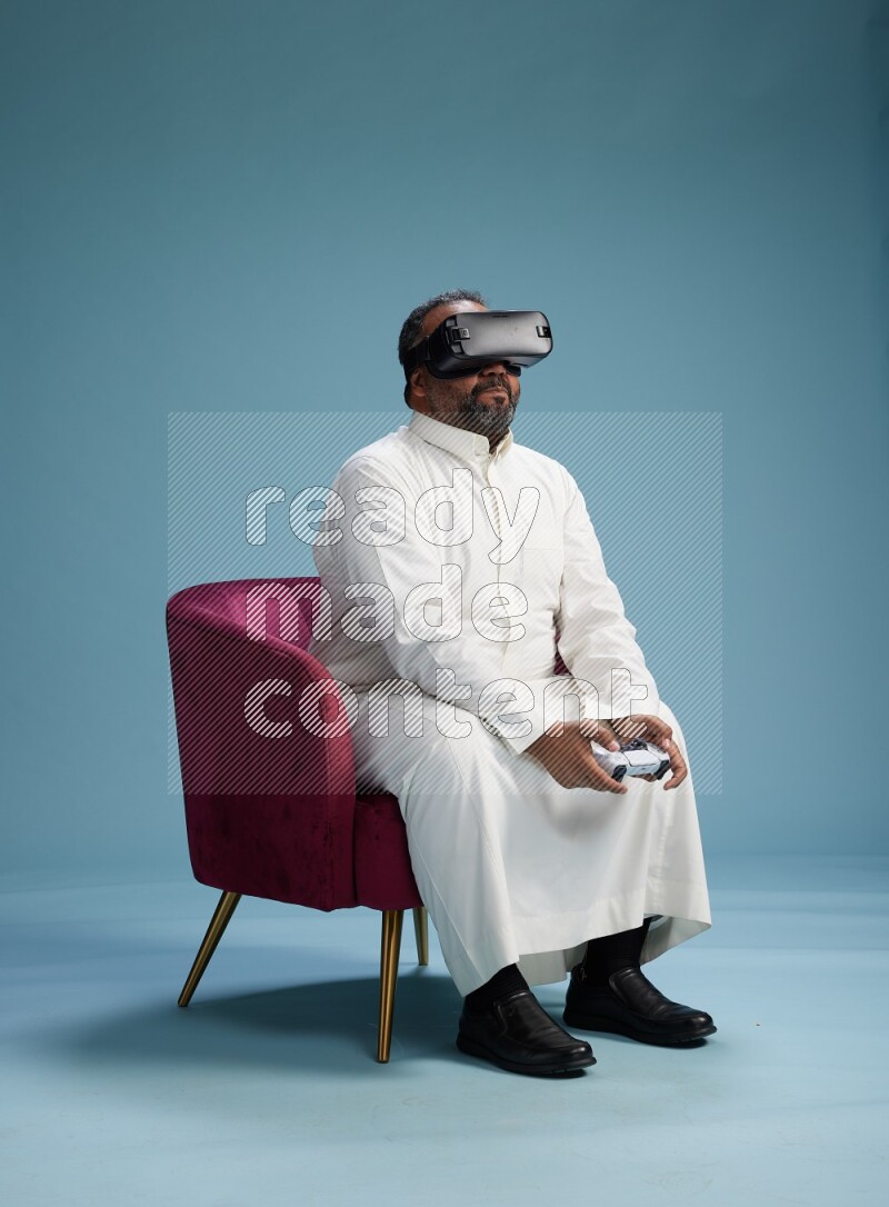 Saudi Man without shimag sitting on chair  gaming with VR and joystick on blue background