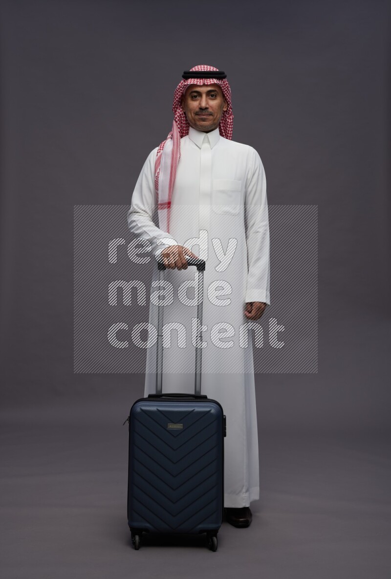 Saudi man wearing thob and shomag standing holding bag on gray background