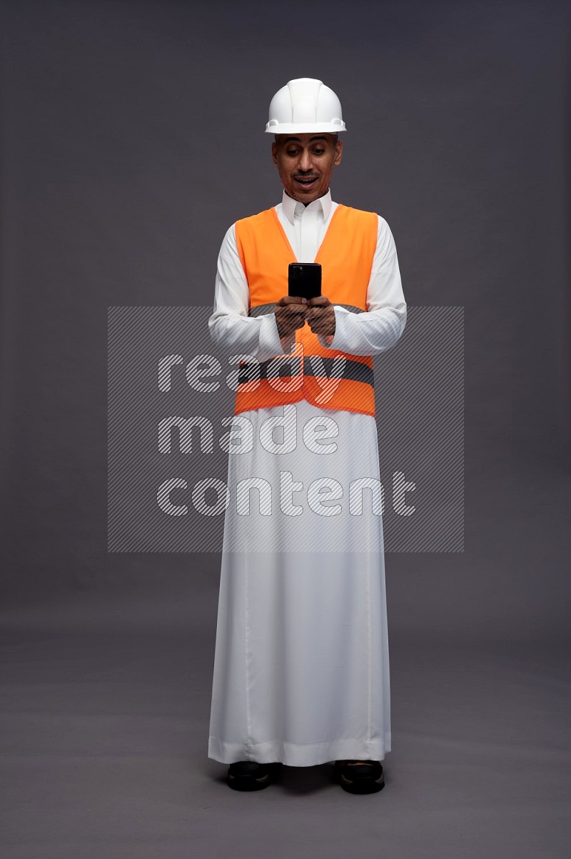 Saudi man wearing thob with engineer vest standing texting on phone on gray background