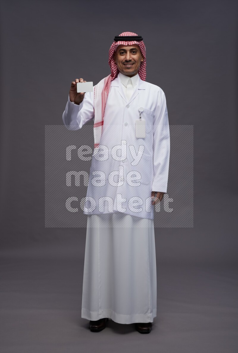 Saudi man wearing thob with lab coat and shomag with pocket employee badge standing holding ATM card on gray background
