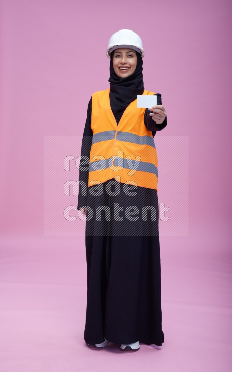Saudi woman wearing Abaya with engineer vest and helmet standing holding ATM card on pink background