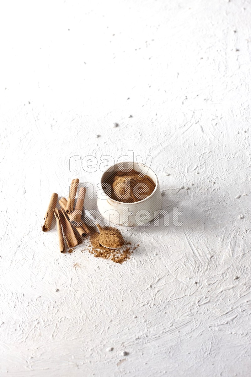 Ceramic beige bowl full of cinnamon powder and a metal spoon with cinnamon sticks next of it on a textured white background