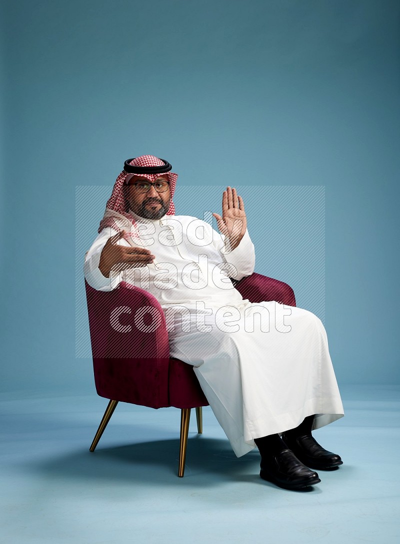 Saudi Man with shimag sitting on chair Interacting with the camera on blue background
