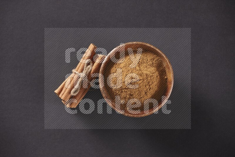 Cinnamon sticks stacked and bounded beside a wooden bowl full of cinnamon powder and a wooden spoon full of powder on black background