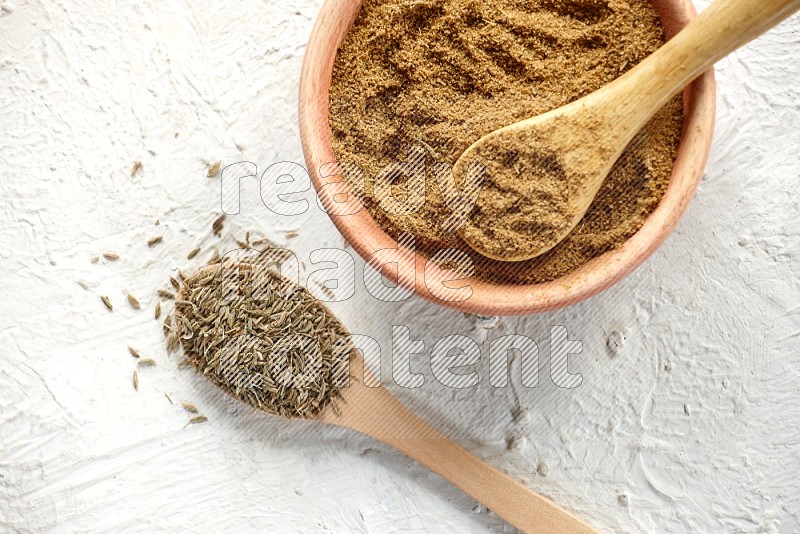A wooden bowl and 2 wooden spoons full of cumin powder and cumin seeds on textured white flooring