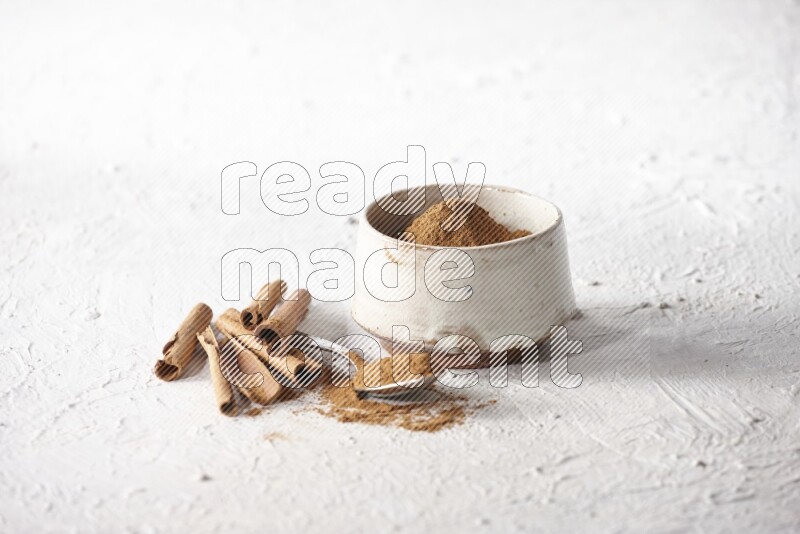 Ceramic beige bowl full of cinnamon powder and a metal spoon with cinnamon sticks next of it on a textured white background