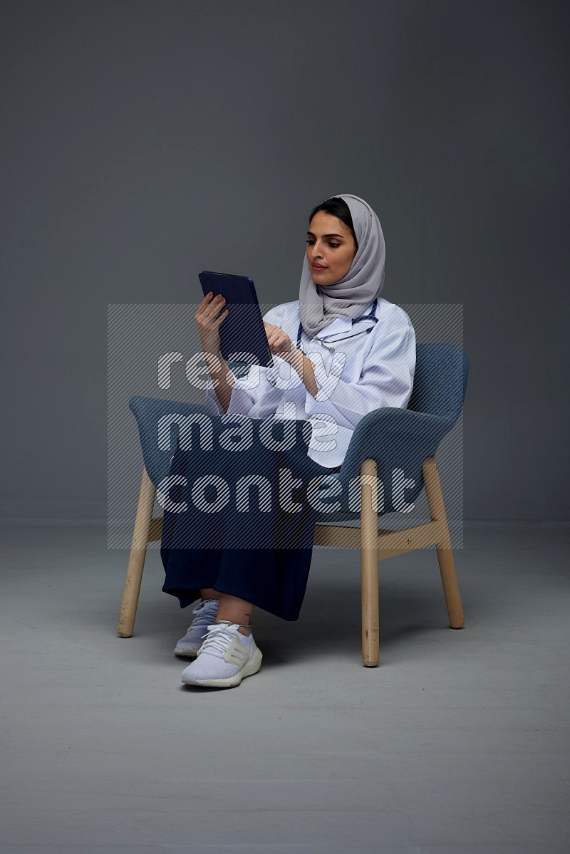 A female doctor wearing a light gray head scarf sitting on a dark grey chair holding an electronic device and pointing to different directions eye level on a grey background