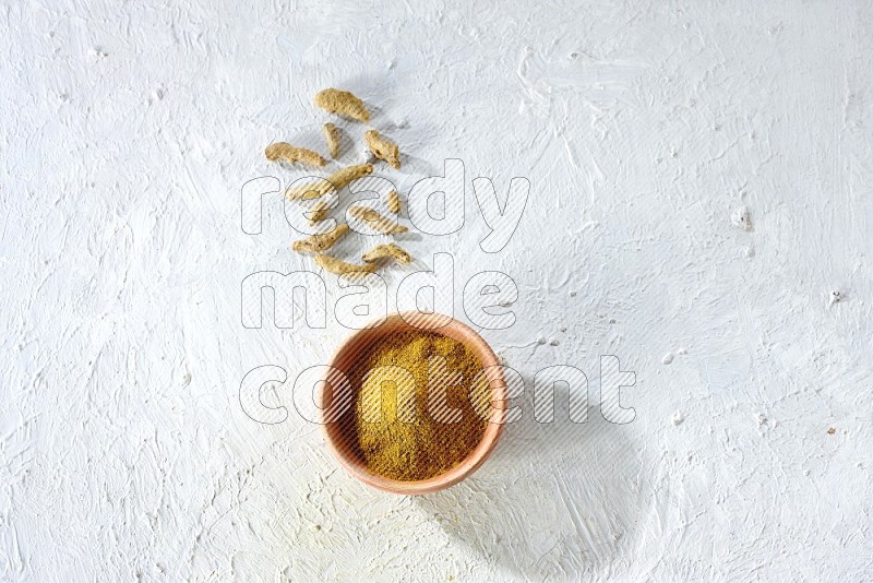 A wooden bowl full of turmeric powder with dried whole fingers on textured white flooring