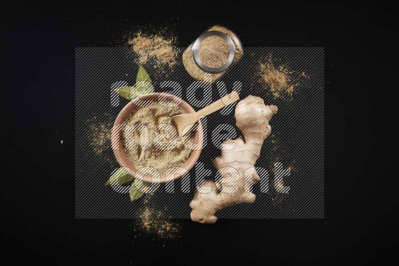 A wooden bowl full of ground ginger powder with a glass jar beside it and fresh ginger on black background