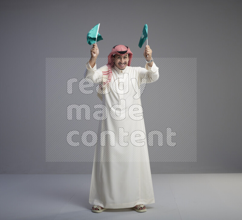 A Saudi man standing wearing thob and red shomag raising small Saudi flag on gray background