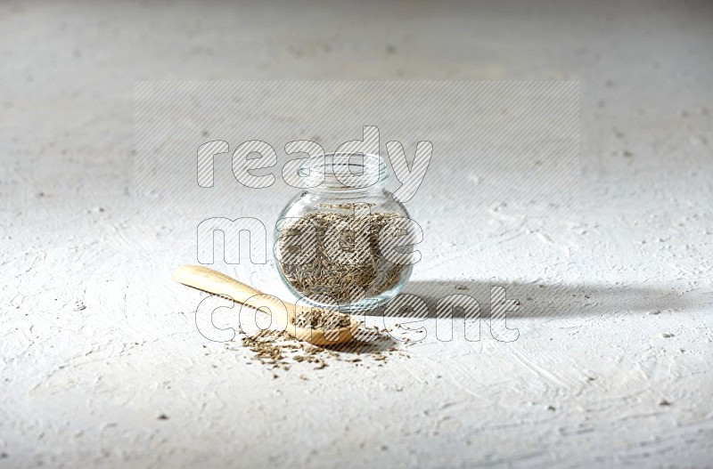 A glass spice jar and wooden spoon full of cumin seeds on textured white flooring