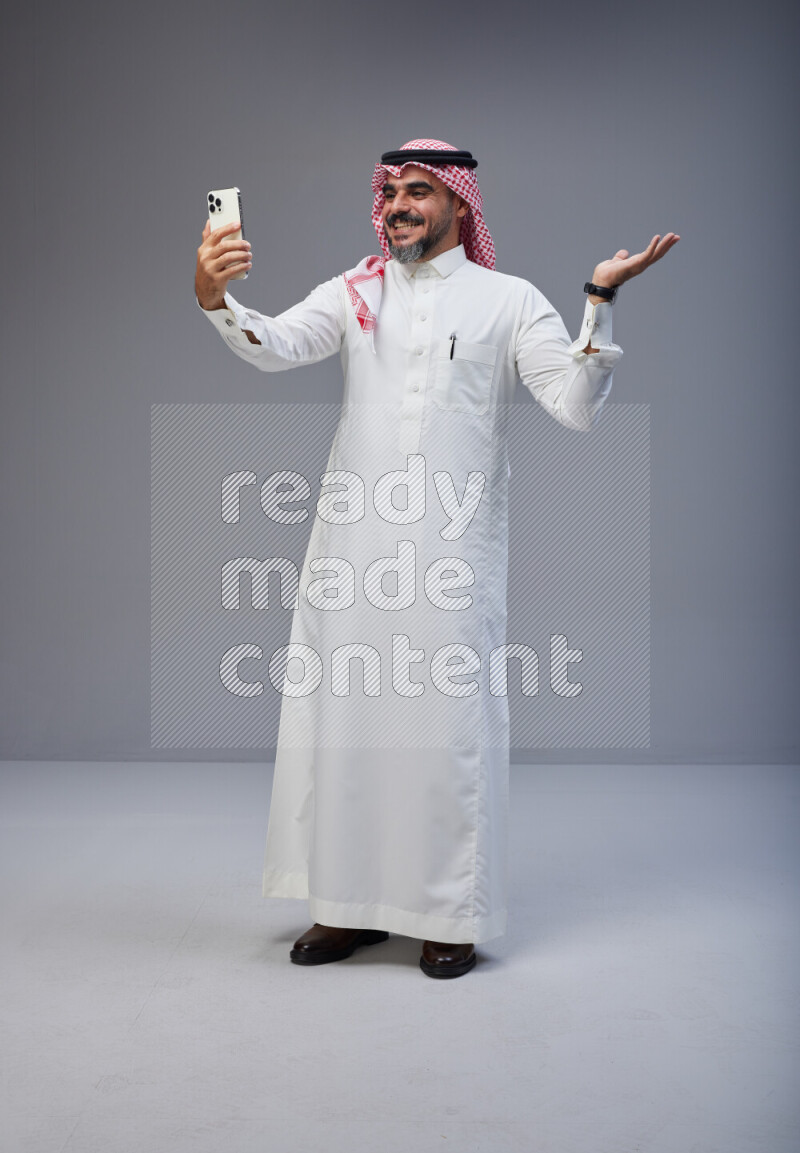 Saudi man Wearing Thob and red Shomag standing taking selfie on Gray background