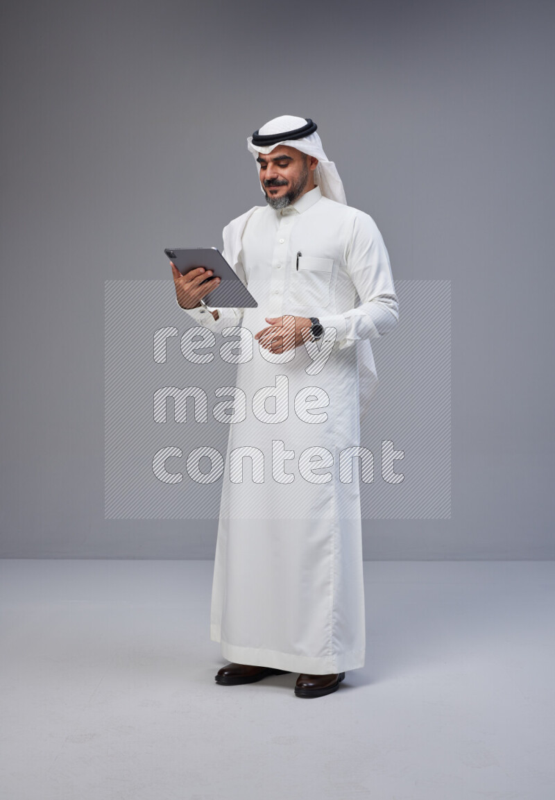 Saudi man Wearing Thob and white Shomag standing working on tablet on Gray background