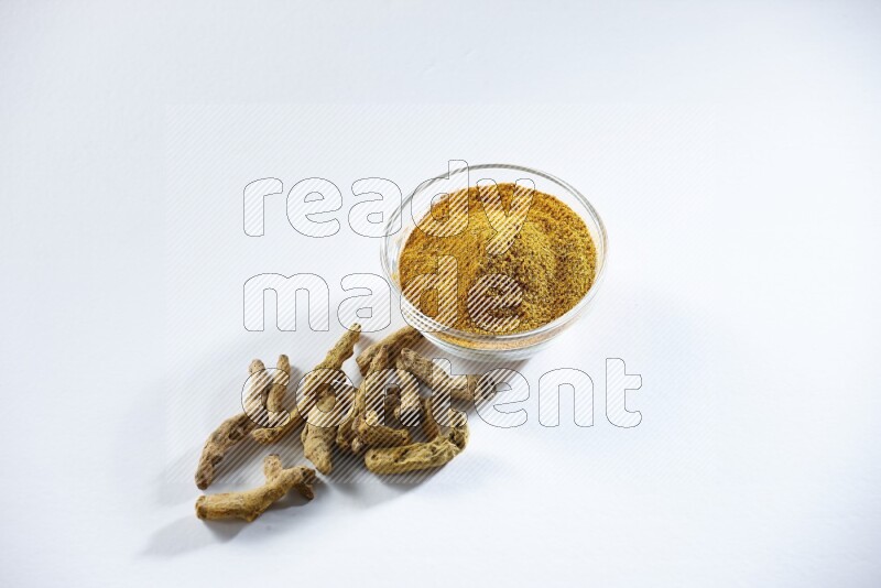 A glass bowl full of turmeric powder and dried whole fingers beneath it on white flooring