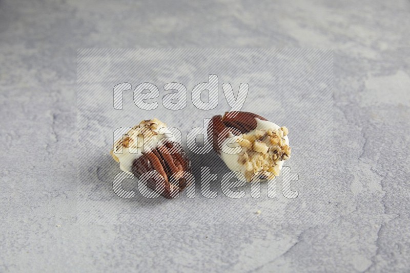 Two pecan stuffed date covered with white chocolate and chopped walnuts on alight grey background
