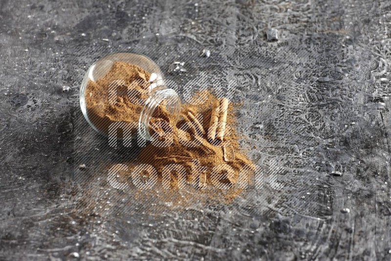 Flipped glass herbs jar full of cinnamon powder with cracked cinnamon sticks on a textured black background