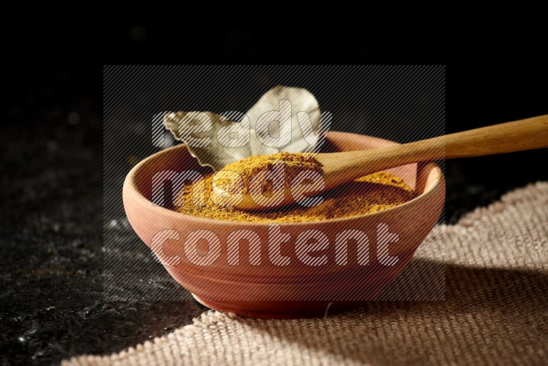 A wooden bowl and a wooden spoon full of turmeric powder on burlap fabric on textured black flooring