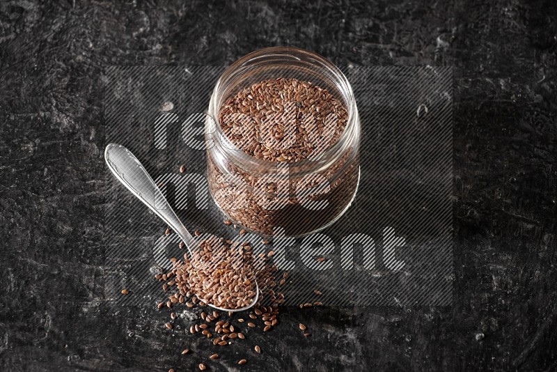 A glass jar full of flaxseeds with a metal spoon full of the seeds on a textured black flooring