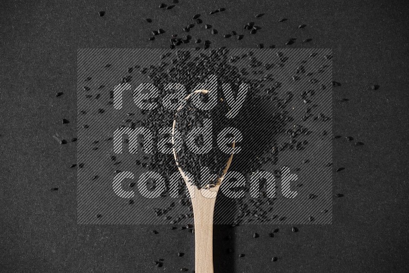 A wooden spoon full of black seeds and surrounded by seeds on a black flooring