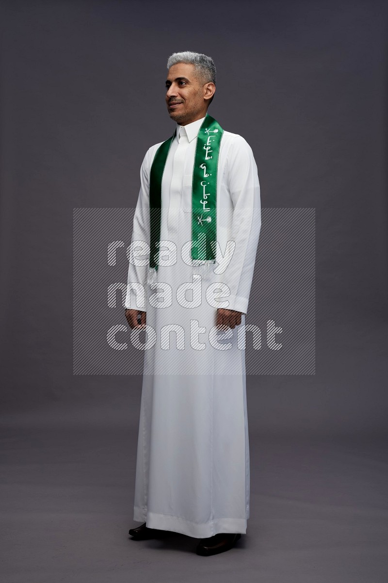 Saudi man wearing thob standing holding Saudi flag on gray background