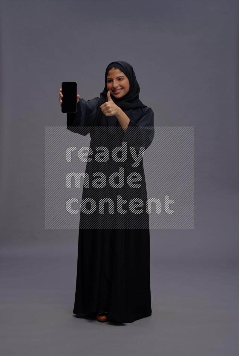 Saudi woman wearing Abaya standing showing phone to camera on gray background