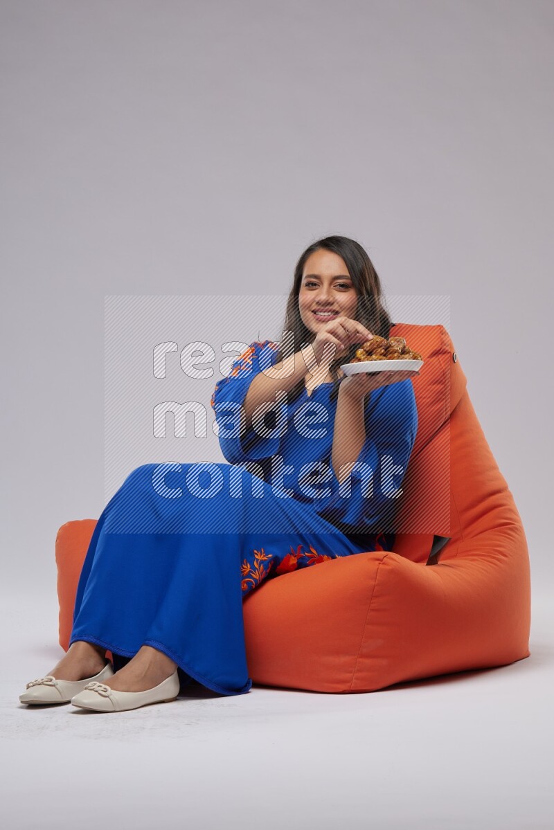 A Woman sitting on an orange beanbag wearing Jalabeya holding a plate of dates