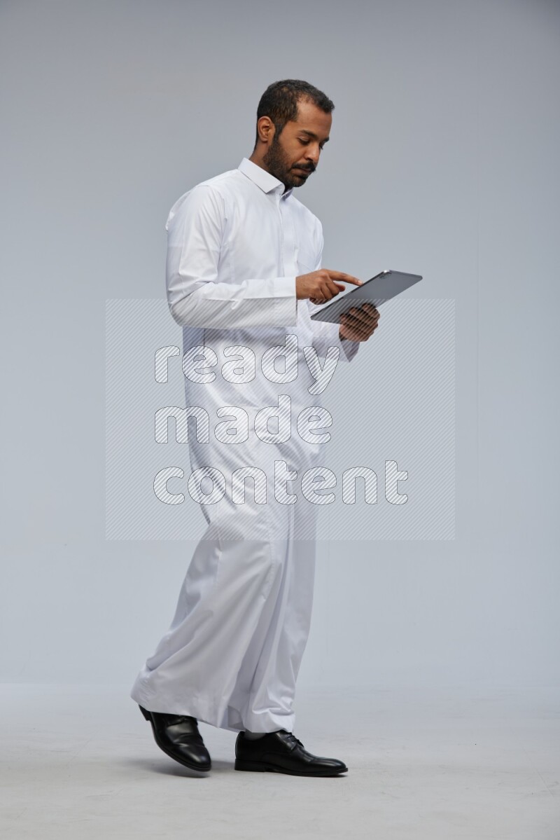 Saudi man wearing Thob standing working on tablet on Gray background