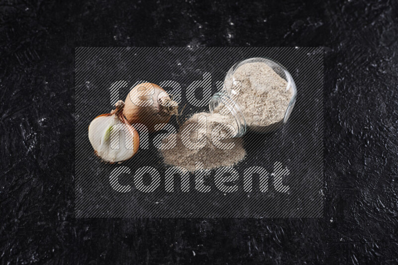 A glass jar full of onion powder flipped with some spilling powder on black background