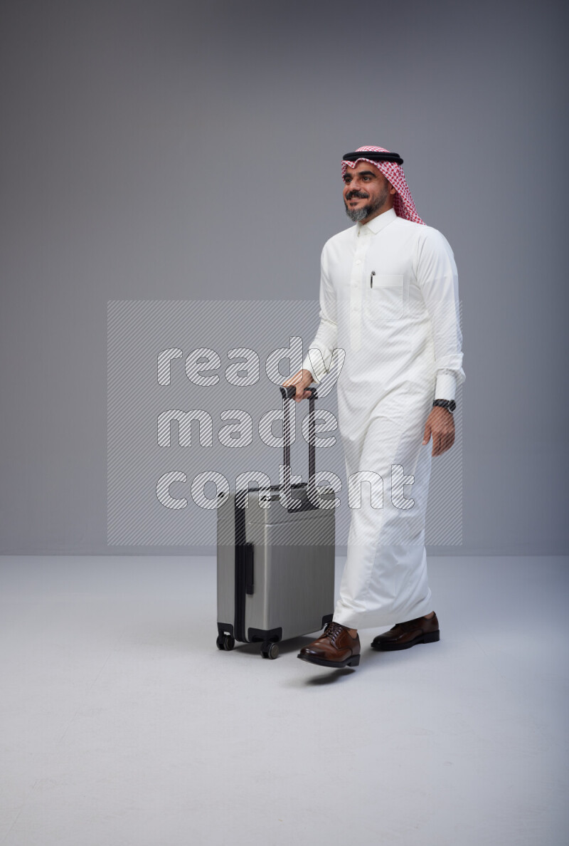 Saudi man wearing Thob and red Shomag standing holding Travel bag on Gray background