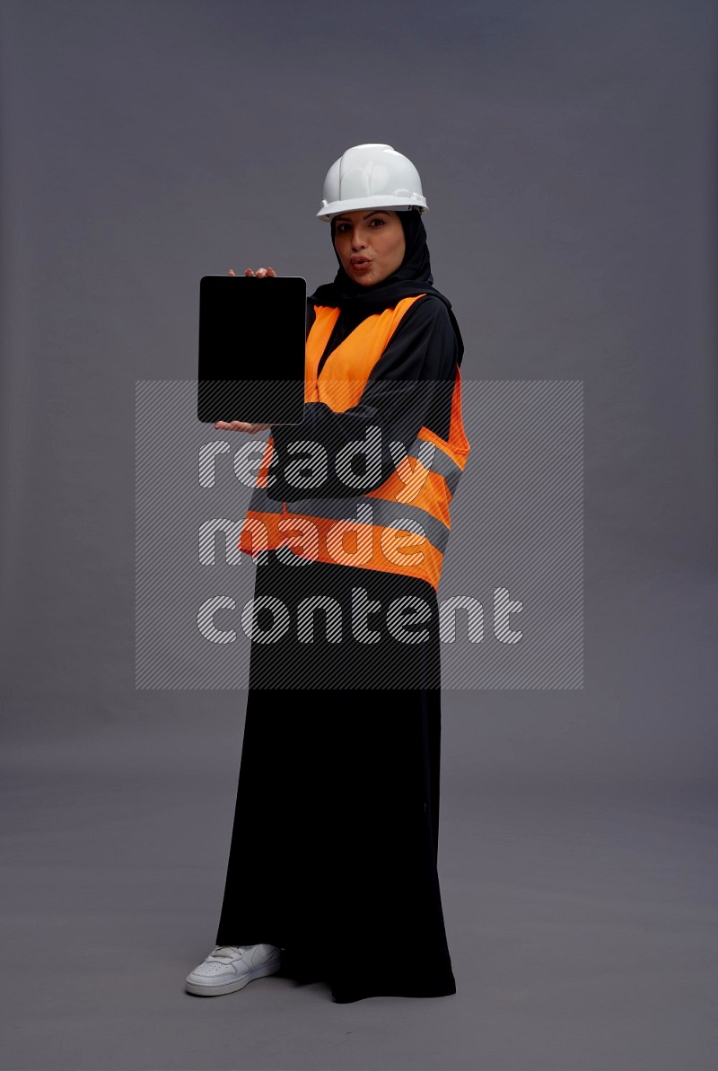 Saudi woman wearing Abaya with engineer vest standing showing tablet to camera on gray background