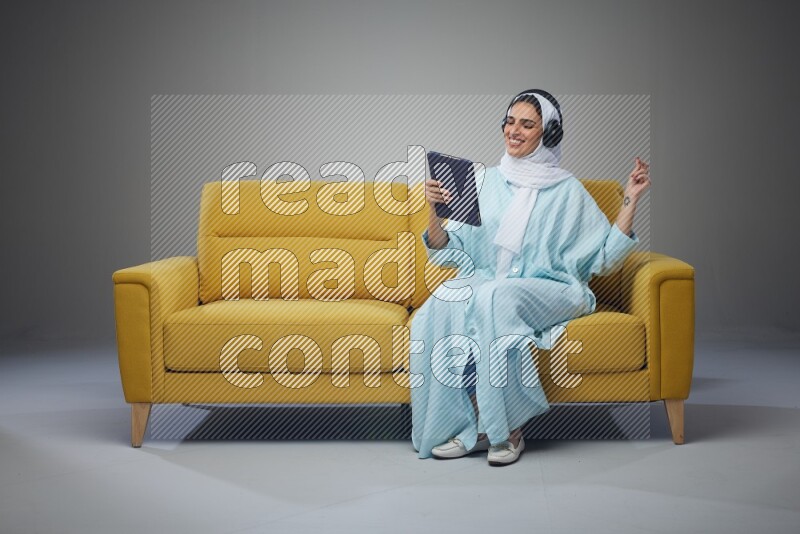 A Saudi woman wearing a light blue Abaya and a white head scarf sitting on a yellow sofa and holding an iPad while wearing headphone eye level on a grey background