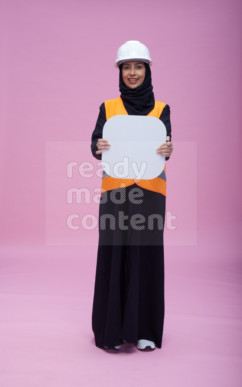 Saudi woman wearing Abaya with engineer vest and helmet standing holding social media sign on pink background