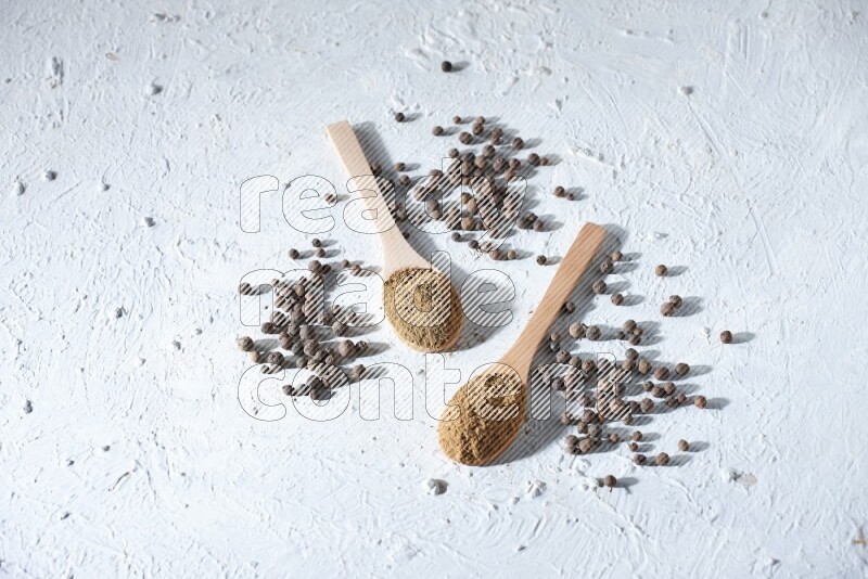 Wooden spoons full of allspice powder and allspice whole balls spreaded on a textured white flooring