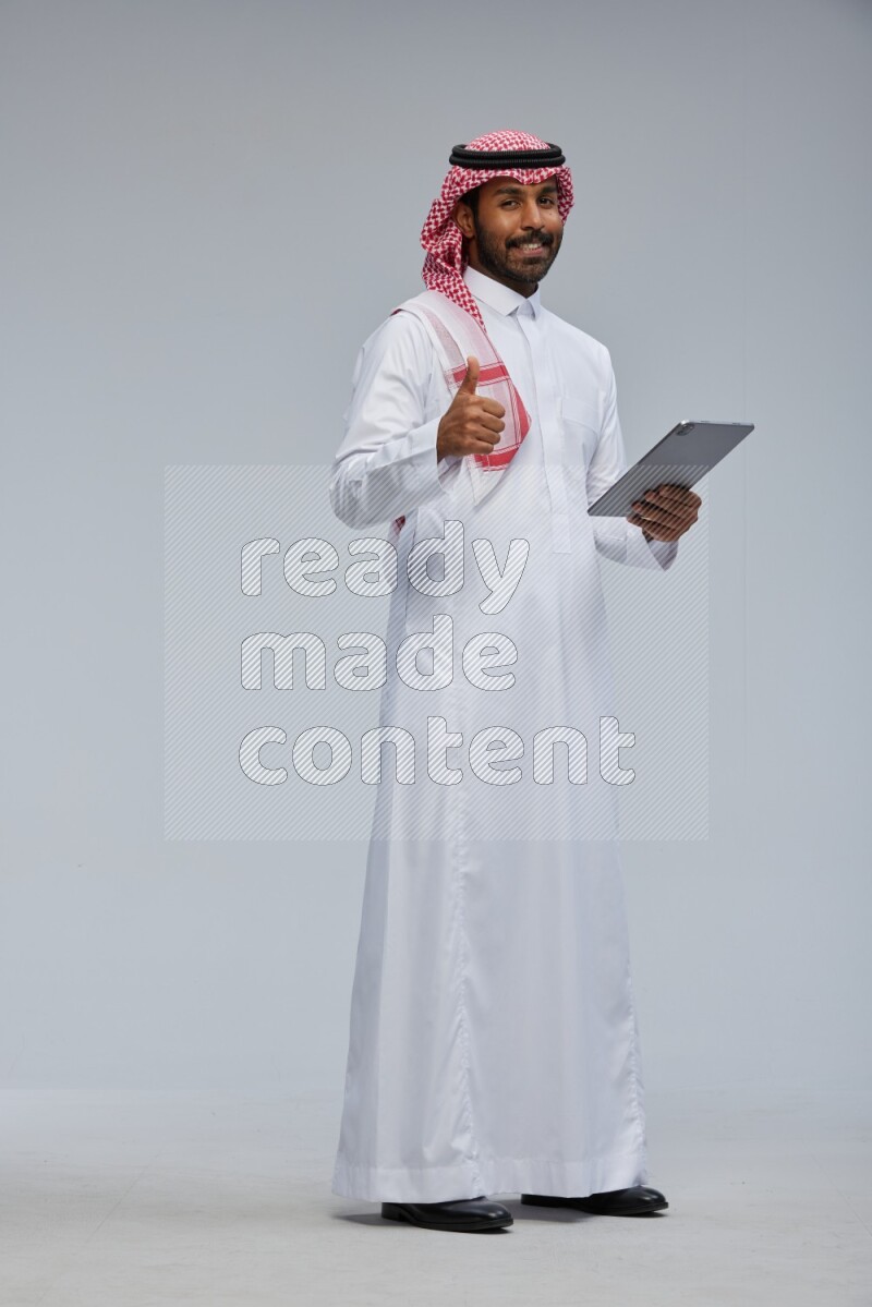 Saudi man Wearing Thob and shomag standing working on tablet on Gray background