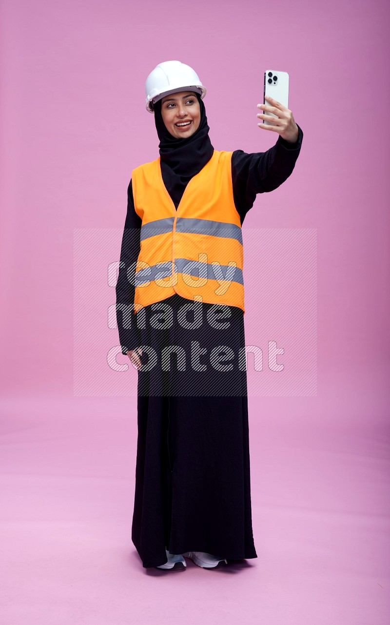 Saudi woman wearing Abaya with engineer vest and helmet standing taking selfie on pink background