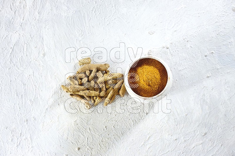 A beige pottery bowl full of turmeric powder and dried turmeric fingers beside it on textured white flooring