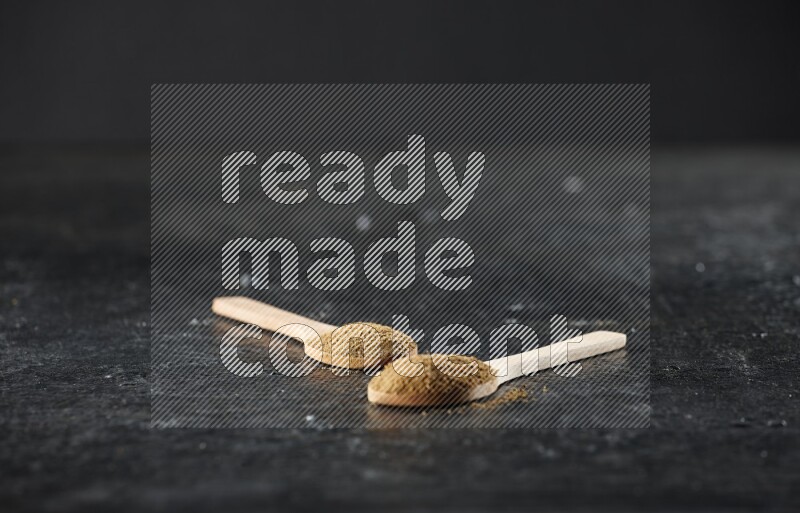 2 wooden spoons full of cumin powder on textured black flooring
