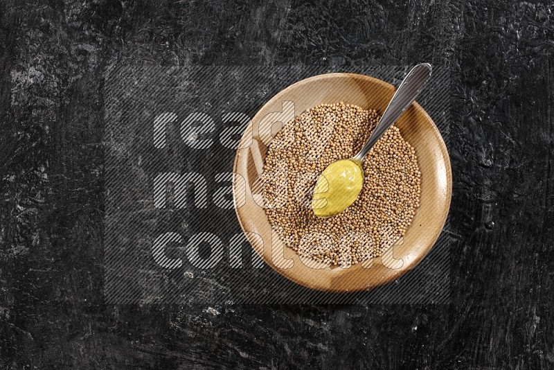 A multicolored pottery plate filled with mustard seeds and a metal spoon full of mustard paste on a textured black flooring in different angles