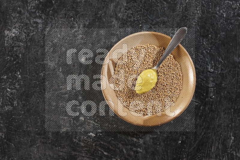 A beige pottery plate filled with mustard seeds with a metal spoon full of mustard paste in it on a textured black flooring