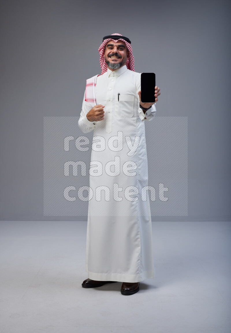 Saudi man Wearing Thob and red Shomag standing showing phone to camera on Gray background